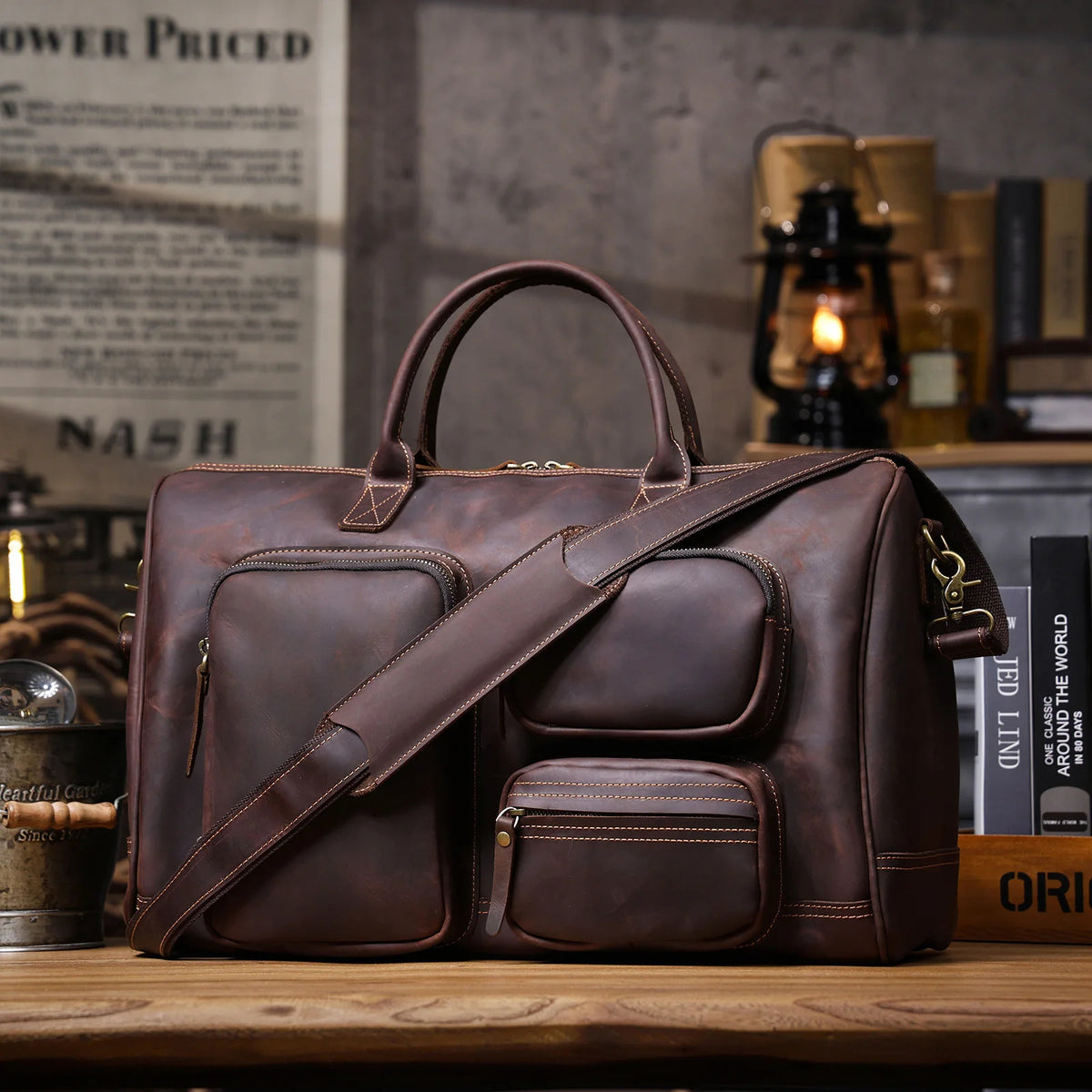 A dark brown leather travel bag with multiple zippered compartments, top handles, and a detachable shoulder strap, displayed on a wooden table in a rustic, vintage-styled room with books and a lantern in the background.