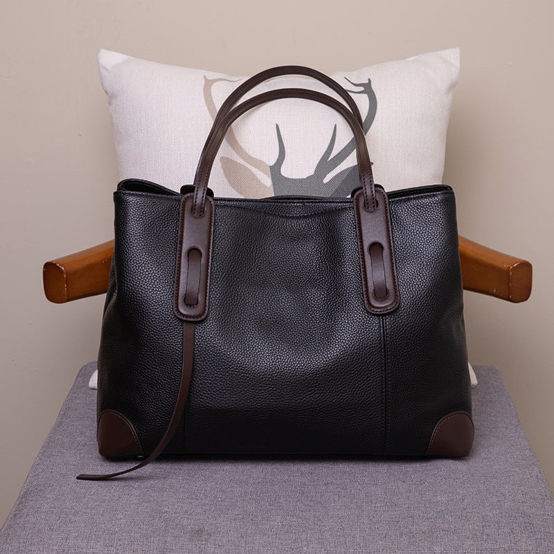 Black leather handbag with brown accents on a gray surface with a white pillow in the background